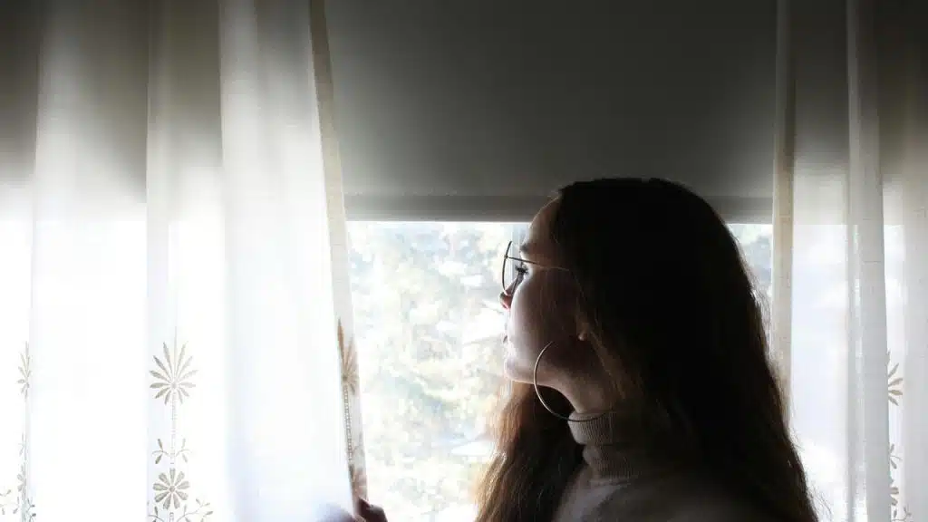 A woman looking out a window through sheer curtains.