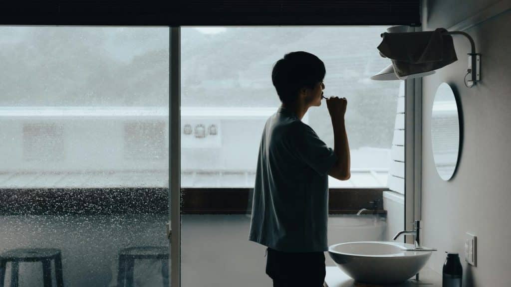 A person brushing their teeth by a sink beside a rainy window.