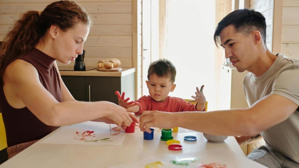 A family painting together at a table, with a child playing with colorful paints.