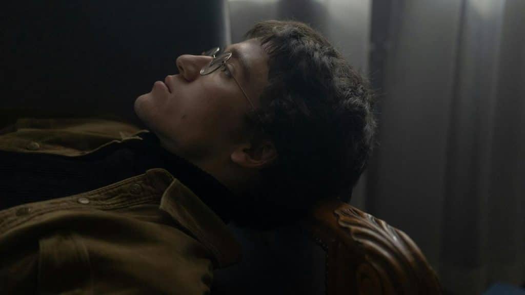 A man wearing glasses leaning back in a chair, gazing upward thoughtfully in dim lighting.