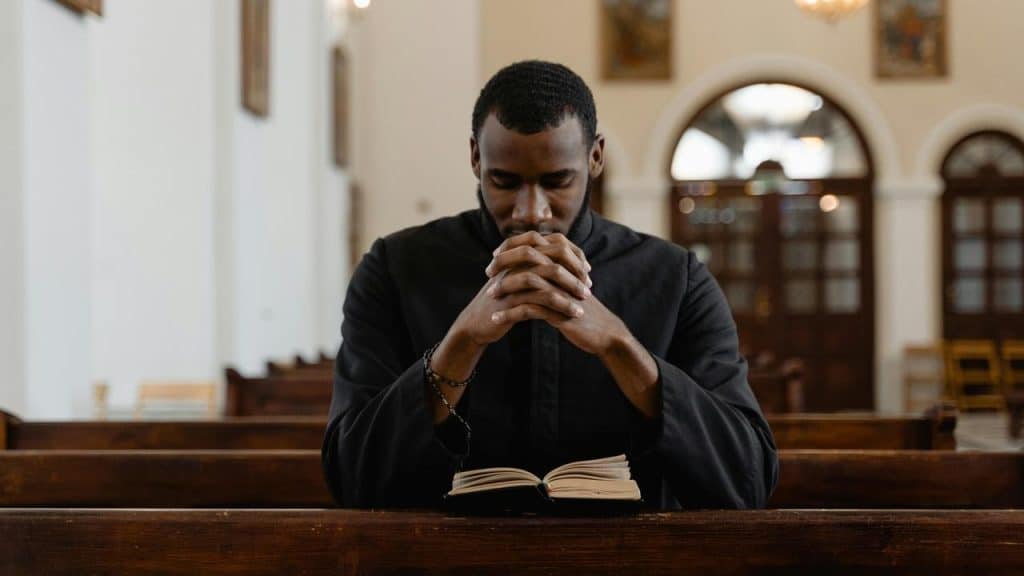 A man sitting in a church pew with hands clasped, praying over an open book.
