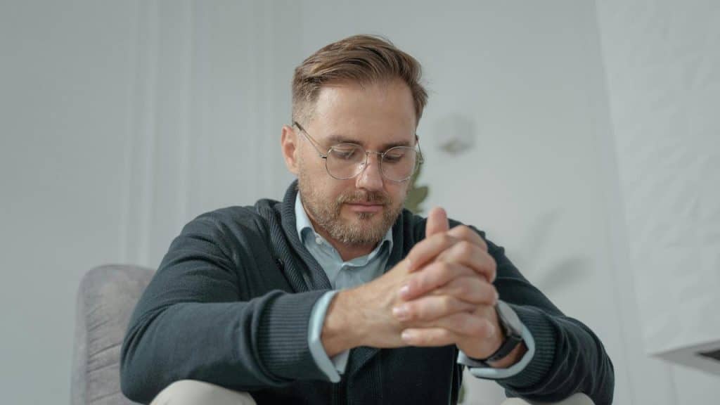 A man with glasses sitting with hands clasped, looking thoughtful.