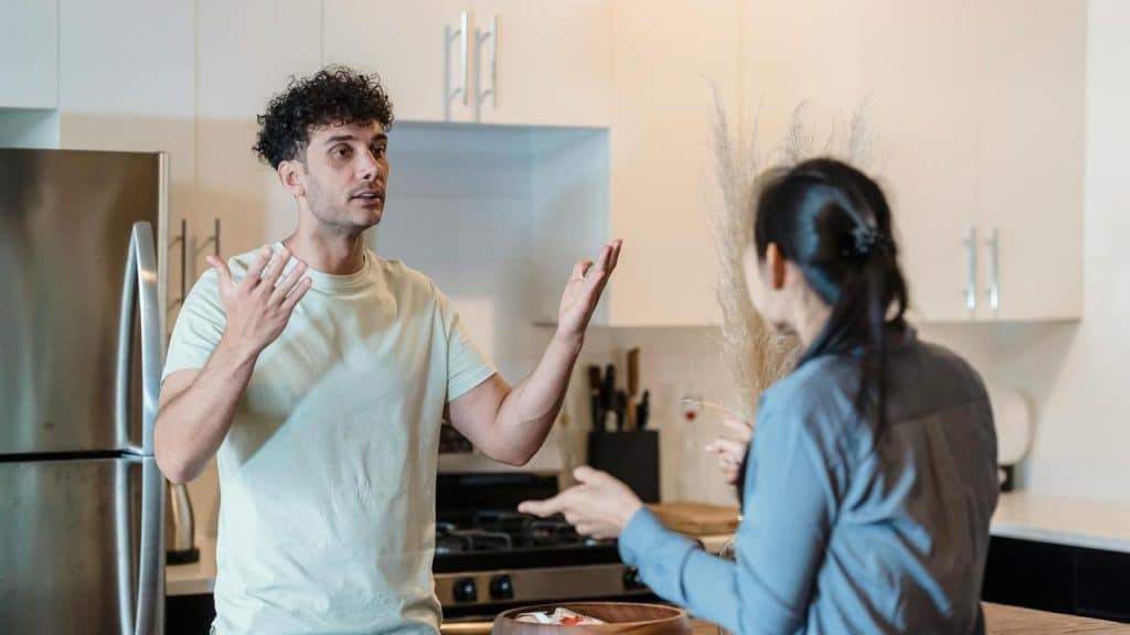 A man and woman arguing in a kitchen, gesturing with their hands.