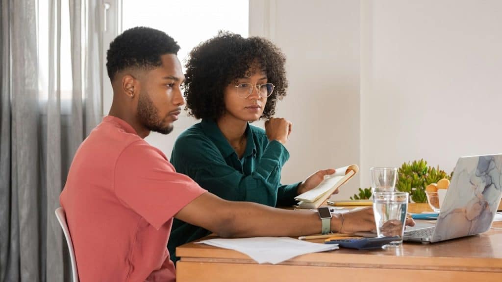 A couple sitting at a table and using a laptop while the woman holds a notepad.