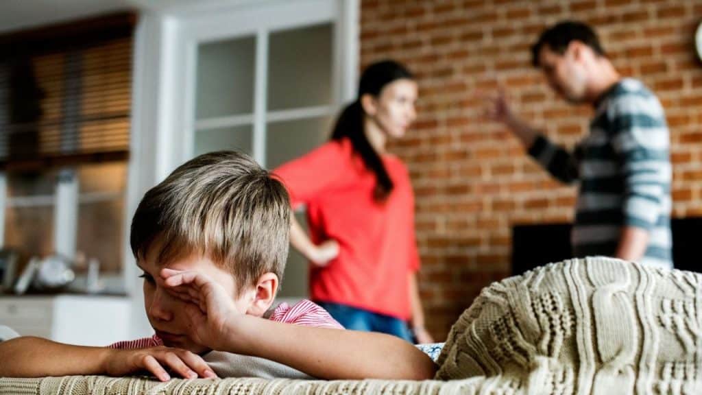 A young boy wipes his eyes while two adults stand blurred in the background.