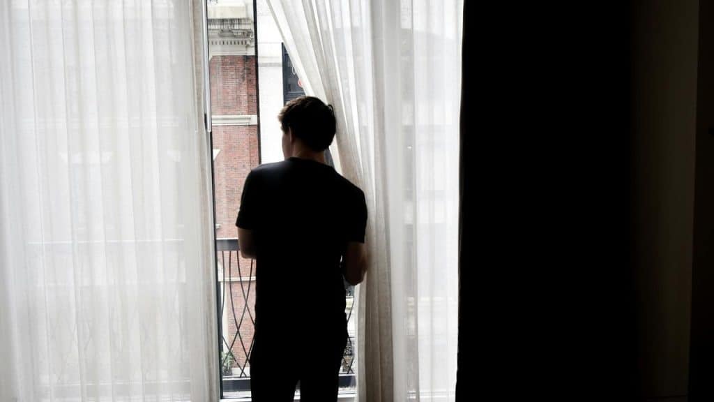 A person standing by a window, looking outside through sheer curtains.