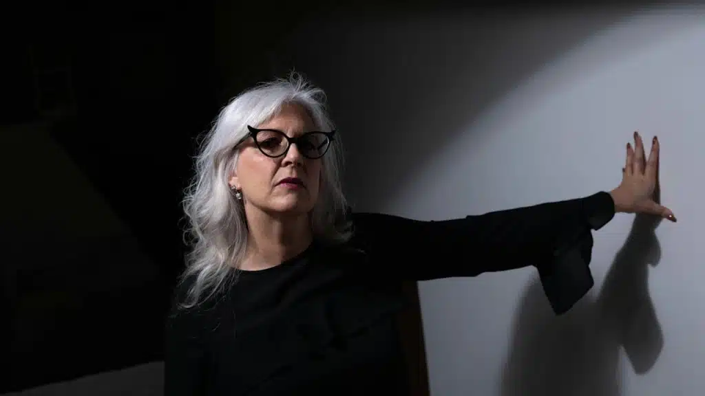 An older woman wearing glasses is leaning on a wall.