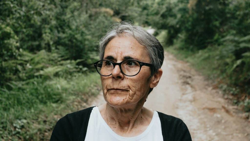 An older woman wearing glasses standing on a walking trek looking distraught.