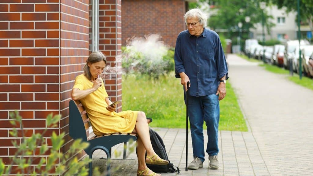 An older woman using a phone and sitting on a bench while ignoring an older man who is looking at her angrily while using a cane.