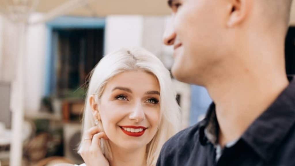 A blonde woman with red lipstick smiles while looking at a man in the foreground.