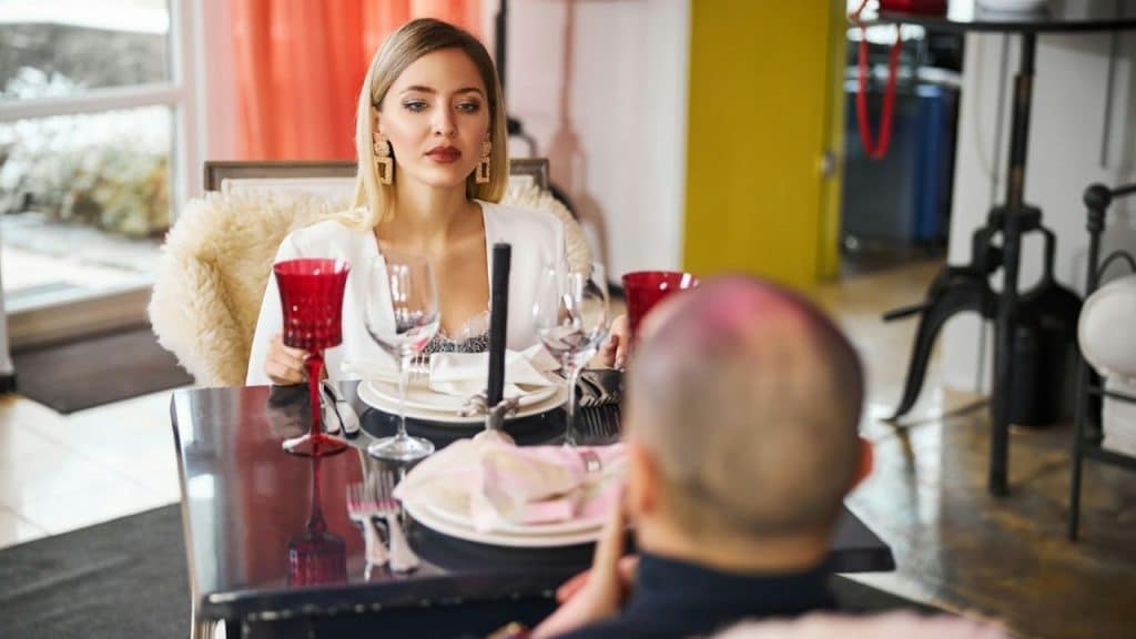 A woman sits at a dinner table with a serious expression, looking at a man.