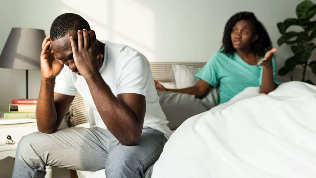 A man sits on a bed with his head in his hands while a woman speaks.