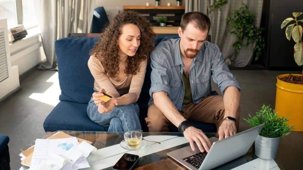 A woman holds a credit card while looking at a man typing on a laptop.