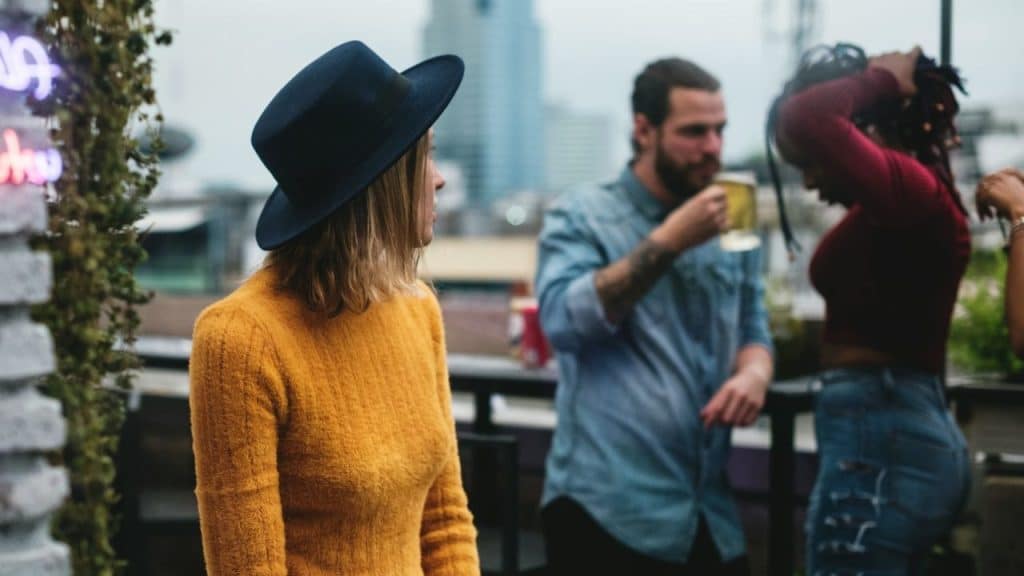 A woman in a yellow sweater and hat looks toward people socializing on a rooftop.