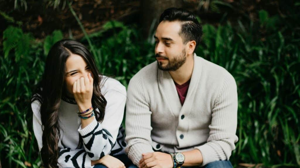 A woman laughs and covers her mouth while a man sits beside her watching.