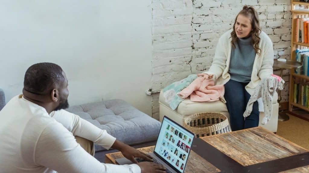 A woman gestures toward baby clothes while a man sits at a laptop nearby.