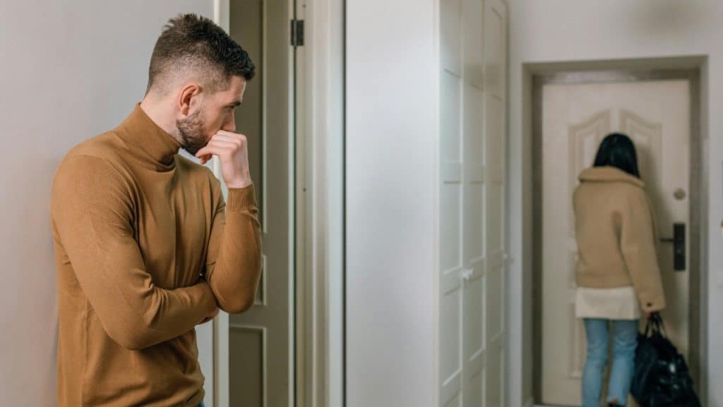 A man watches a woman from a hallway as she walks toward a door.
