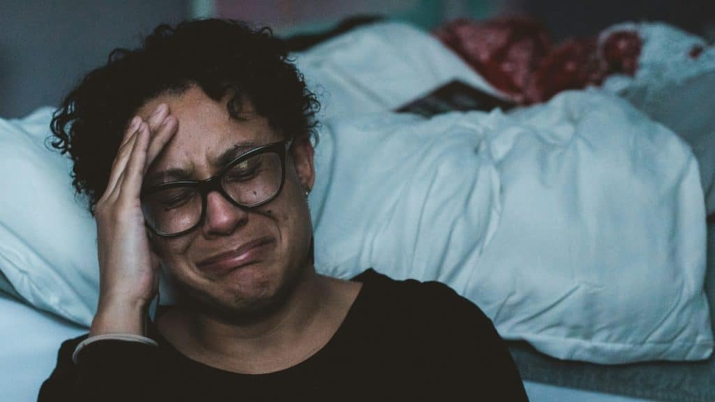 A person wearing glasses with curly hair crying while leaning against white bedding in low light.