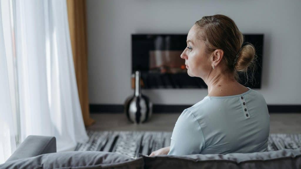 A woman with blonde hair in a bun sitting on a couch, looking toward a window.