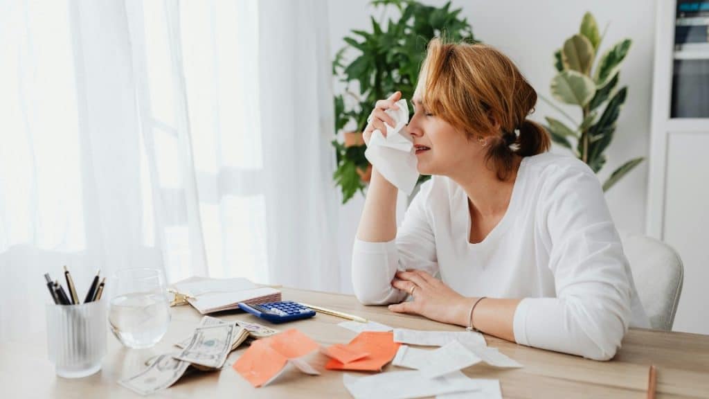 A woman at a desk crying into a tissue with bills, money, and a calculator nearby.
