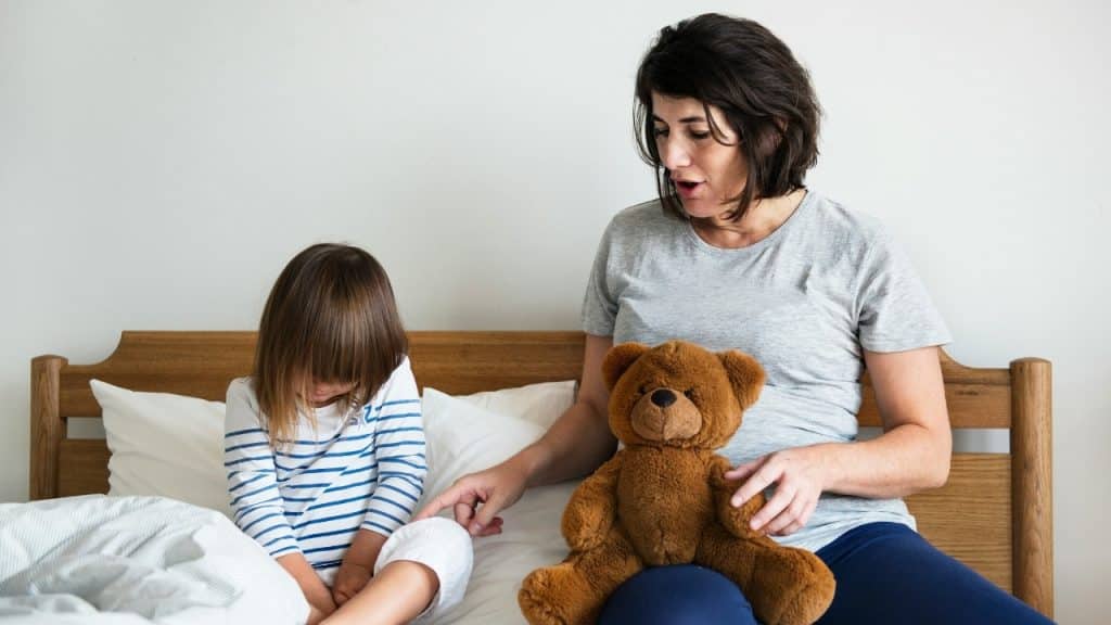 A woman sitting on a bed next to a child and a brown teddy bear, gesturing.