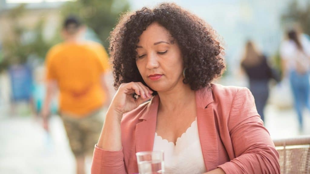 A woman with curly hair looking down with her hand on her chin at an outdoor table.