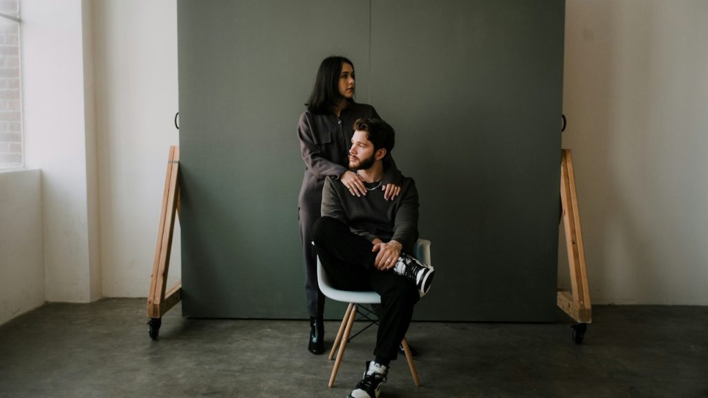 A woman resting her hands on a man’s shoulders as he sits on a chair while she sits on its armrest.
