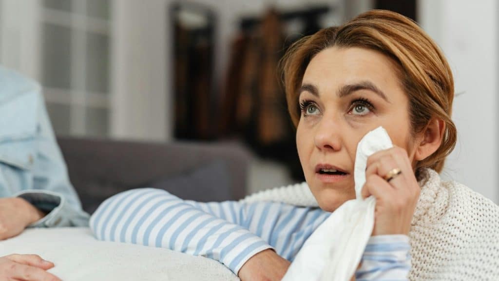 A woman with light brown hair looks upward while holding a white tissue to her face.
