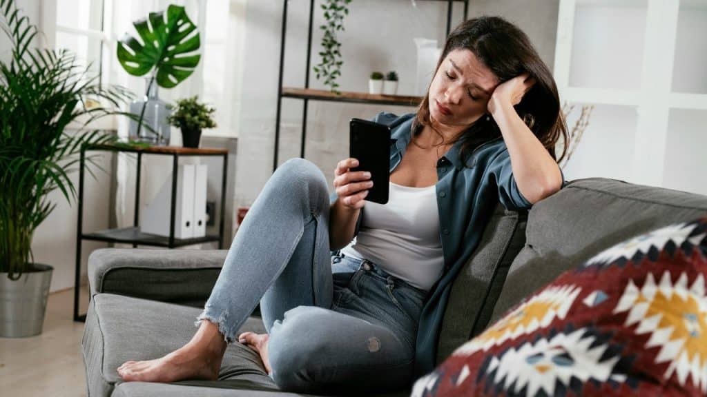 A woman sits on a couch leaning her head on her hand while looking at a phone.