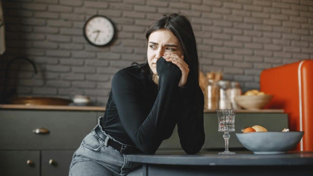 A woman in black leaning on a kitchen table with a somber expression near a bowl.