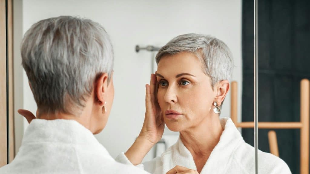 A woman with short gray hair looking at her reflection in a mirror while touching her face.