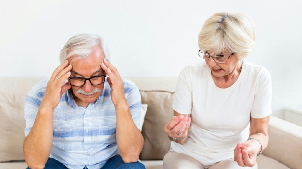An older man holds his head while an older woman speaks and gestures beside him.
