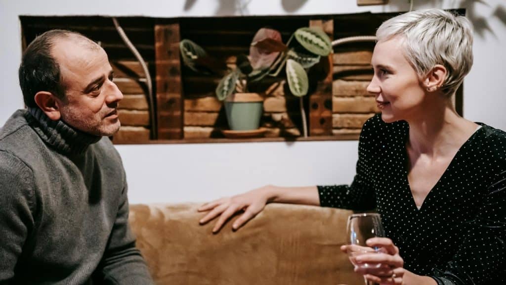 A man in a sweater and a woman with short hair talk on a couch.