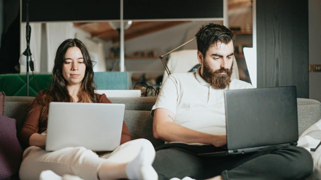 A man and a woman sit together on a sofa, each using their own laptop.