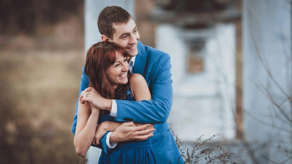 A man in a blue suit hugs a woman from behind while both are smiling brightly.