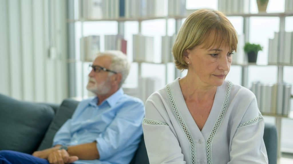 A woman looks down pensively while a man sits out of focus in the background.