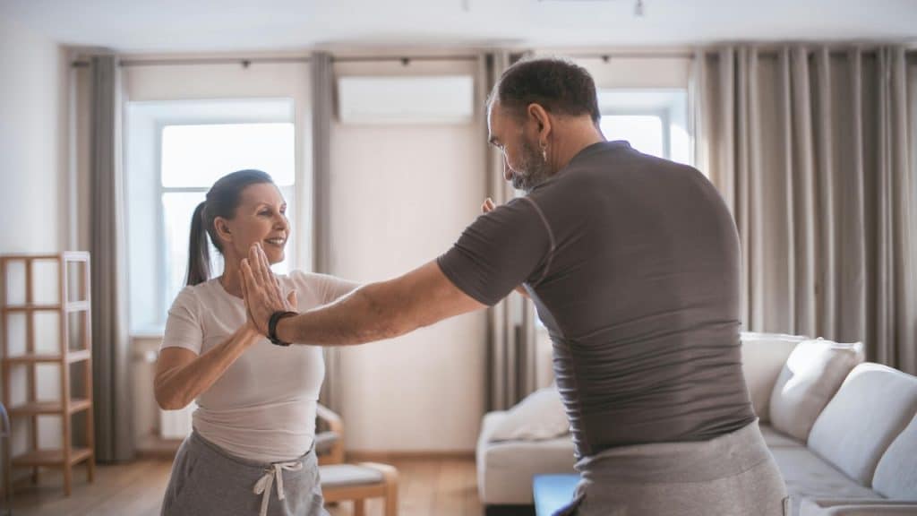 A woman and man in athletic wear touch palms while exercising in a living room.