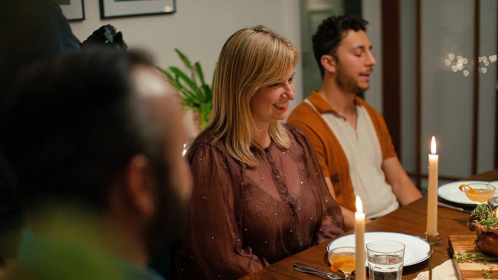 A woman smiles at a dinner table set with candles while other people sit nearby.