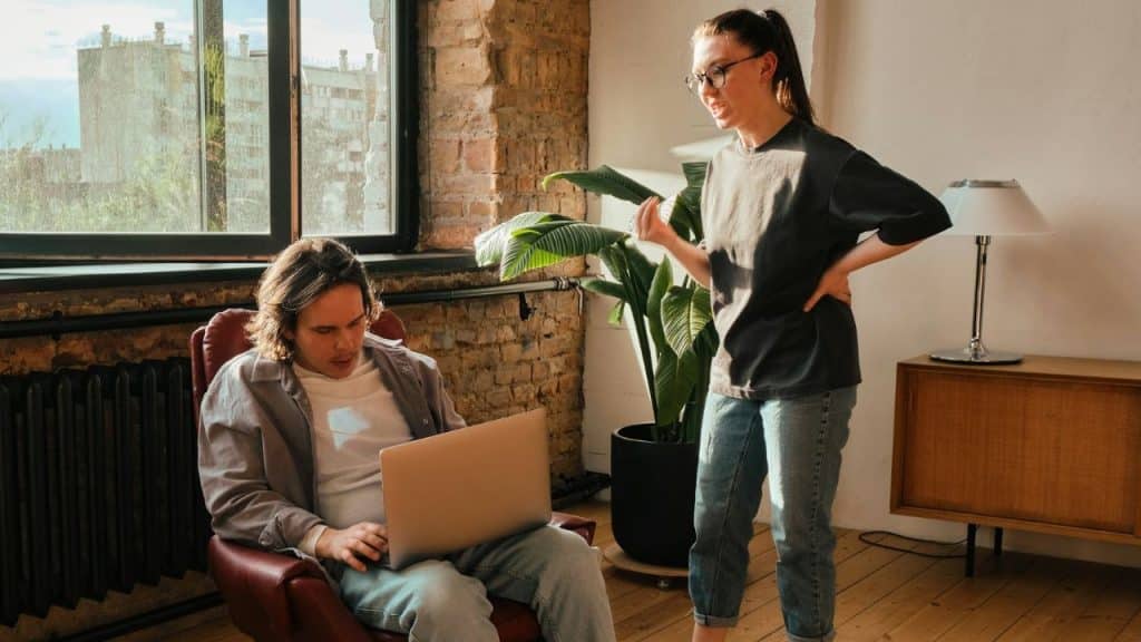 A woman stands and speaks animatedly to a man who is looking at a laptop.