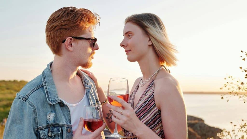 A couple holding glasses of rosé wine and gazing at each other by the water at sunset.