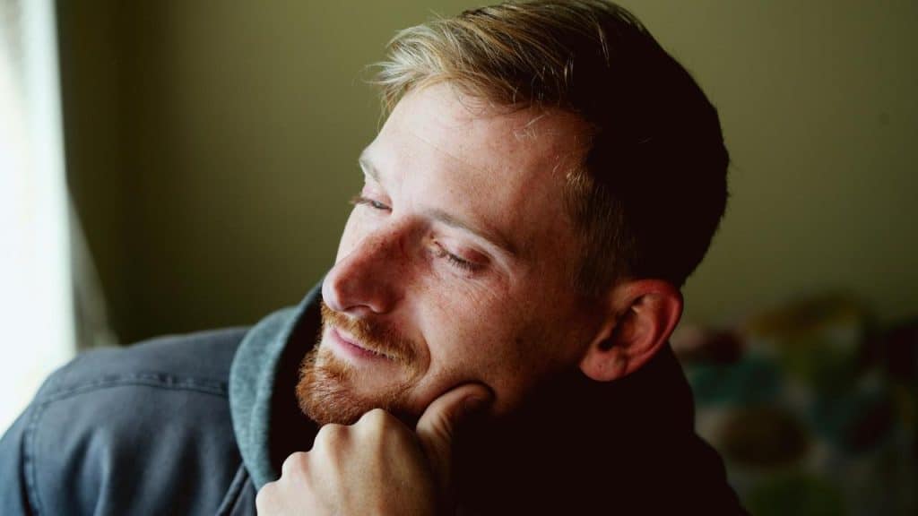 A man with a thoughtful smile resting his chin on his hand by a window.