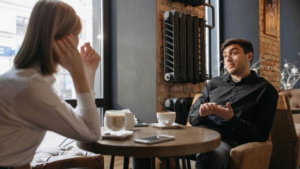 A man and woman talking across a table in a café with coffee cups.