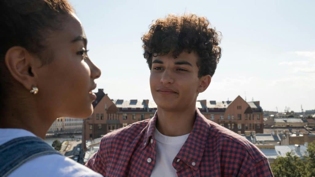 A man and woman talking outdoors with a cityscape in the background.