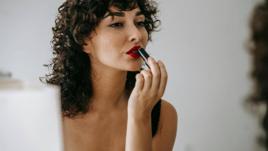 A person with curly hair applying red lipstick while looking into a mirror.