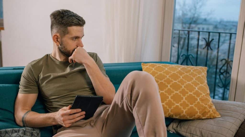 A man sitting on a couch holding a tablet and looking out a window thoughtfully.