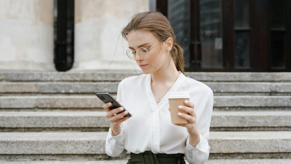 A woman holding a coffee and looking at her phone while standing on steps.