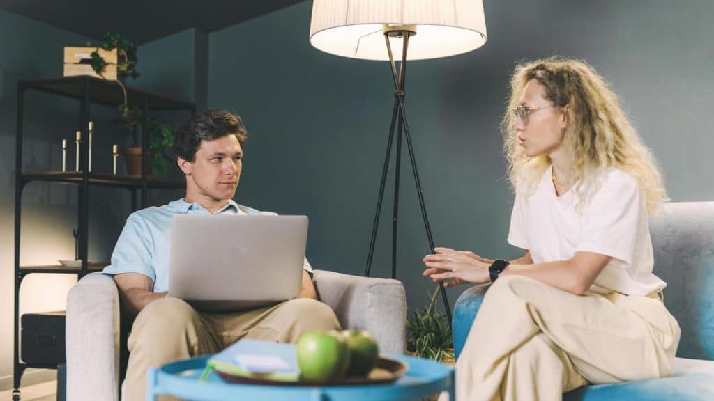 A woman talking to a man using a laptop while sitting in a living room.