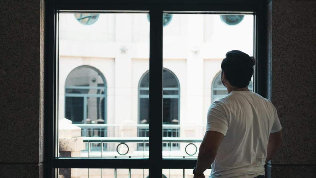 A man standing indoors looking out through a large window.