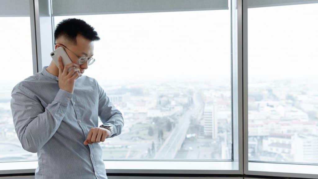 A man talking on the phone while checking his watch by a window.