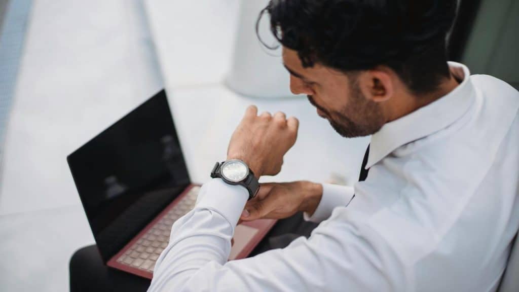 A man checking his watch while sitting with a laptop.